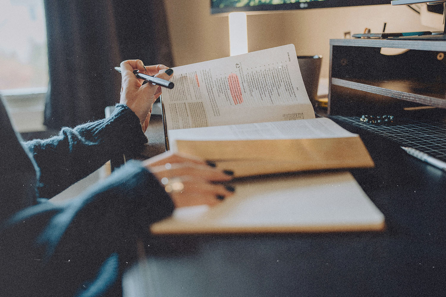 A person studies at a desk with an open Bible, highlighting passages while writing notes in a notebook