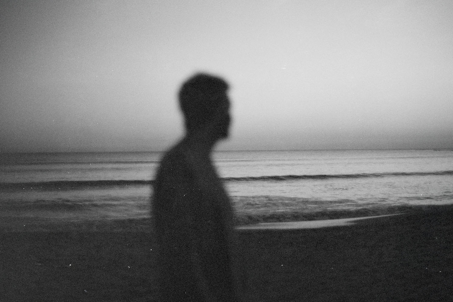 A silhouetted person stands near the shoreline at dusk, with calm waves and a fading horizon behind them