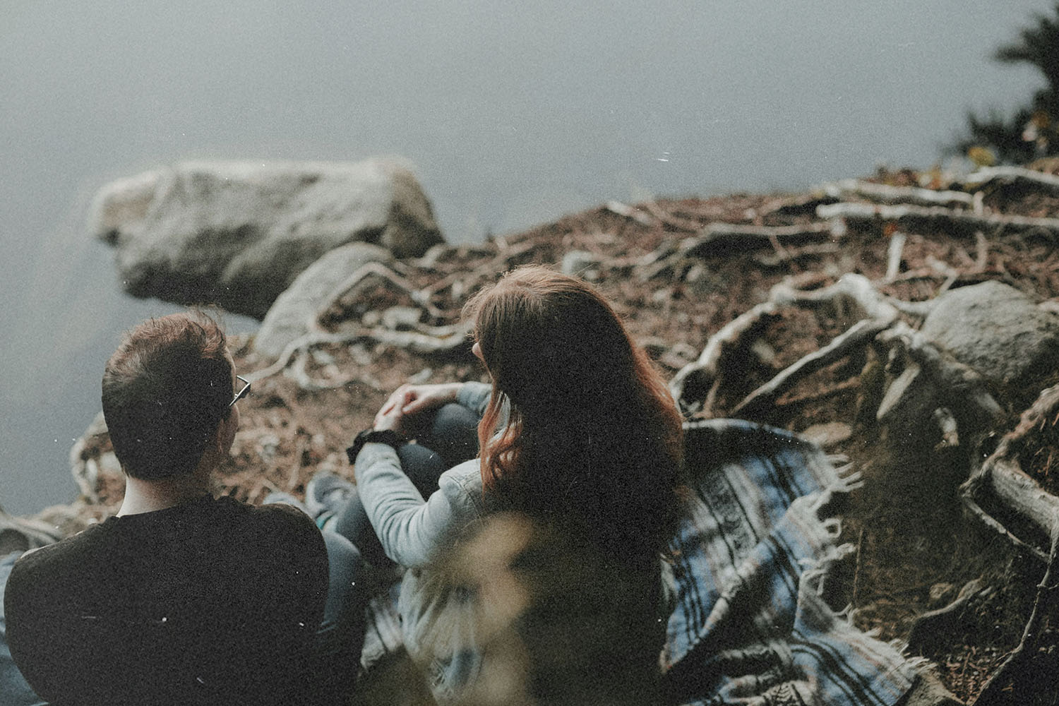 Two people sit on a blanket beside a calm lake, surrounded by tree roots and rocks along the shoreline