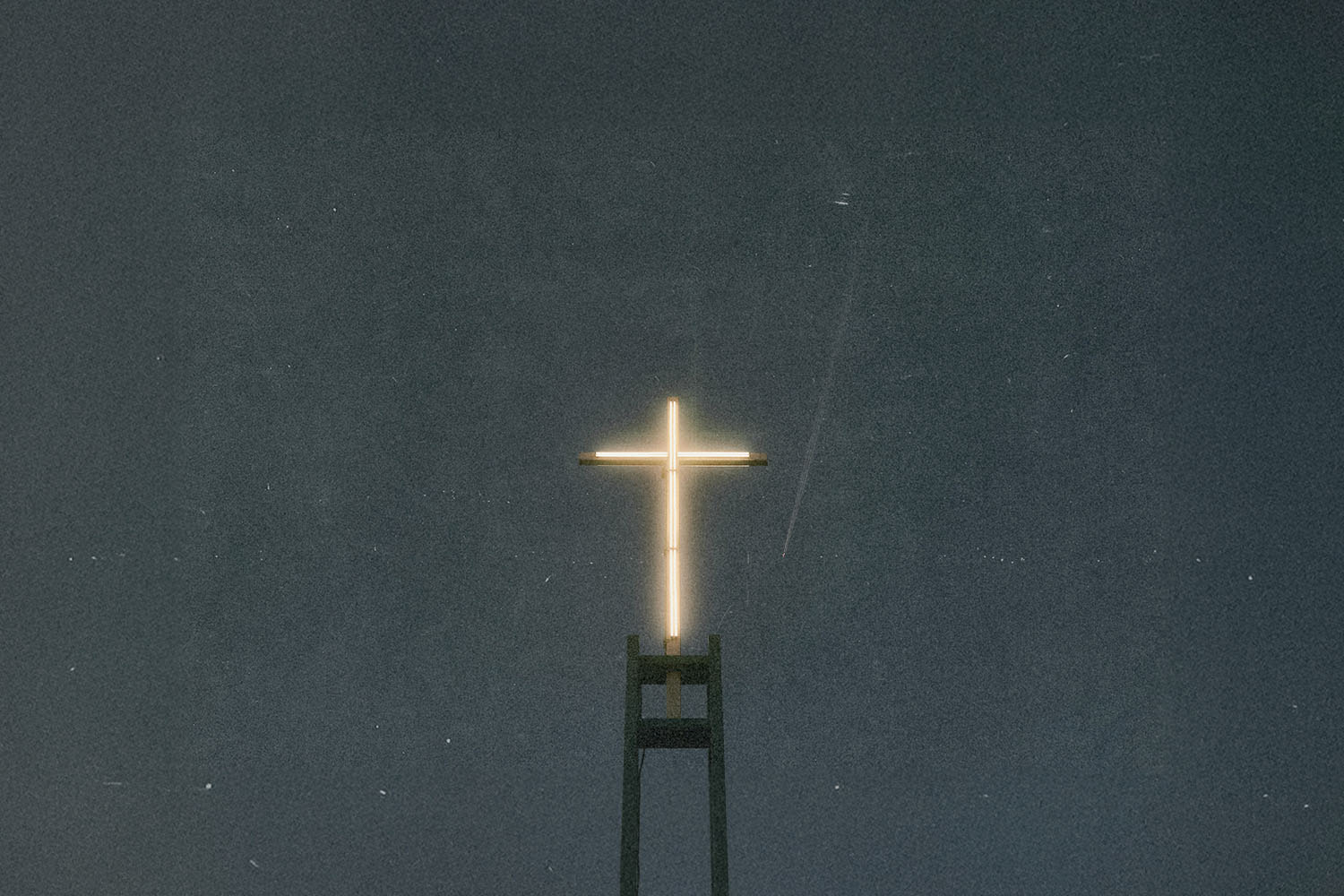 A glowing cross sits atop a tall structure against a dark, starless night sky