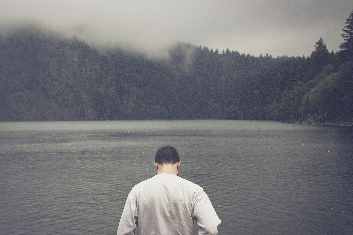 A person stands at the edge of a calm lake surrounded by misty, forested mountains