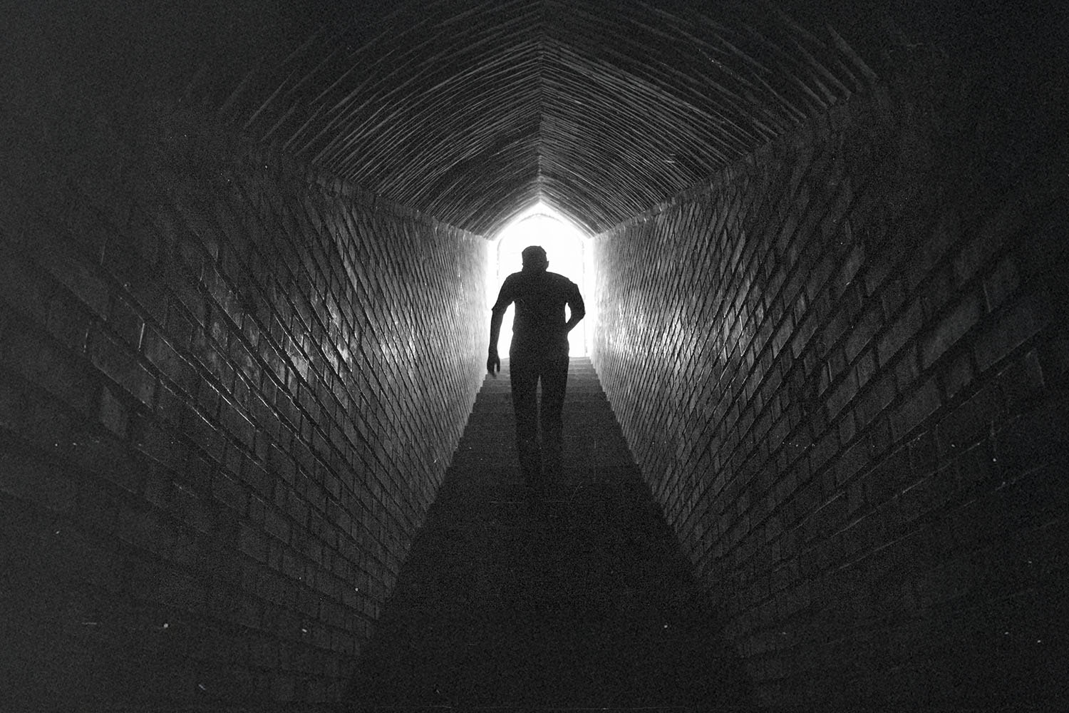 A person walks through a long, dark brick tunnel toward a bright opening at the far end
