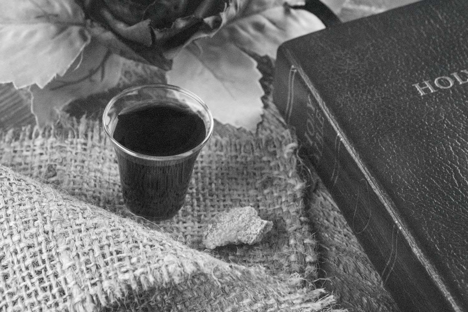 A small cup of communion juice sits on a piece of burlap alongside a broken piece of bread, with a Bible and decorative leaves arranged nearby in a softly lit setting