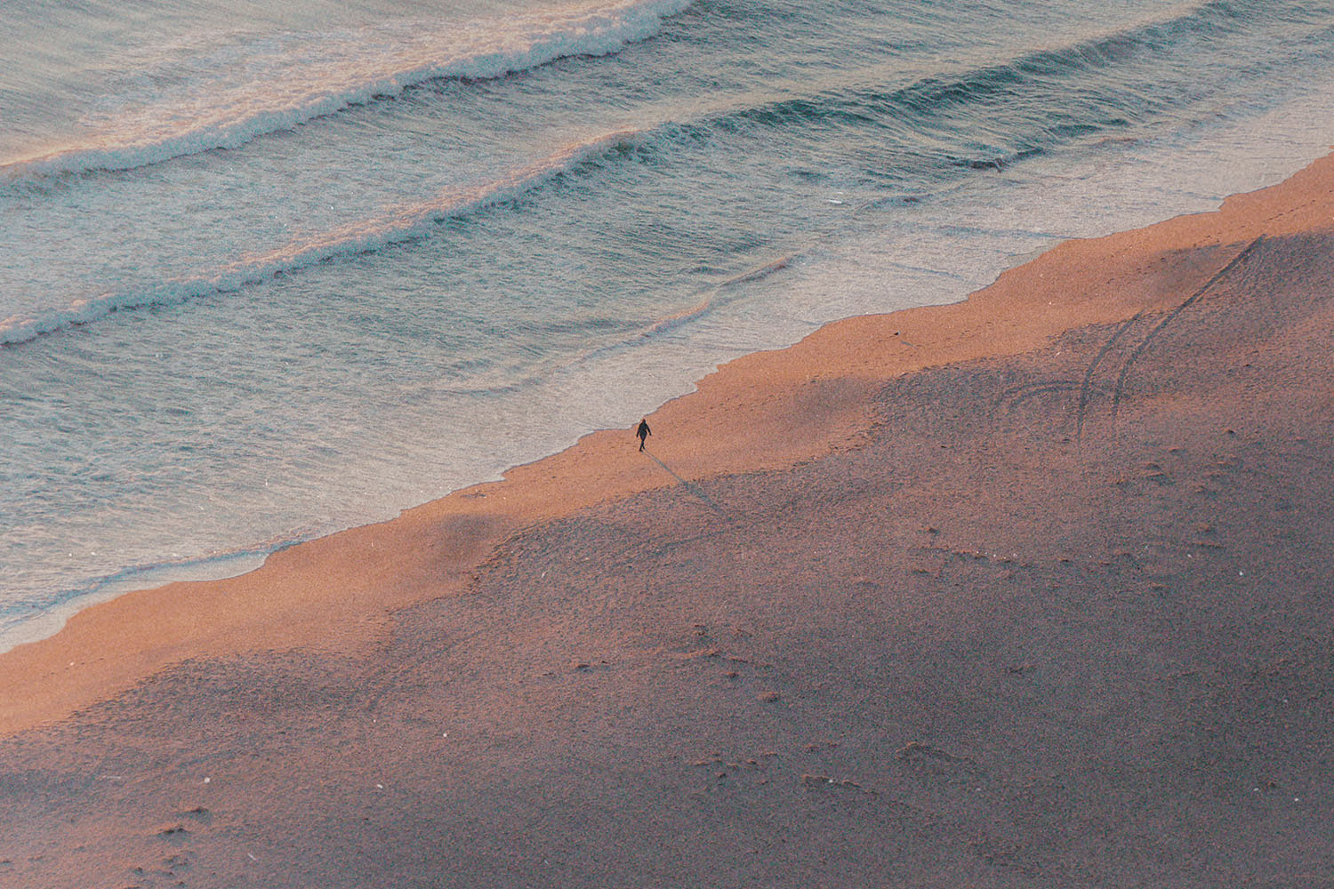 A single person walks along the shoreline at sunrise or sunset, casting a long shadow across the sand as gentle waves roll in