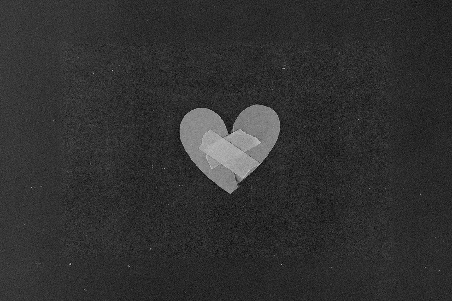 A simple paper heart sits against a dark background with two pieces of tape placed in an “X” shape across the center