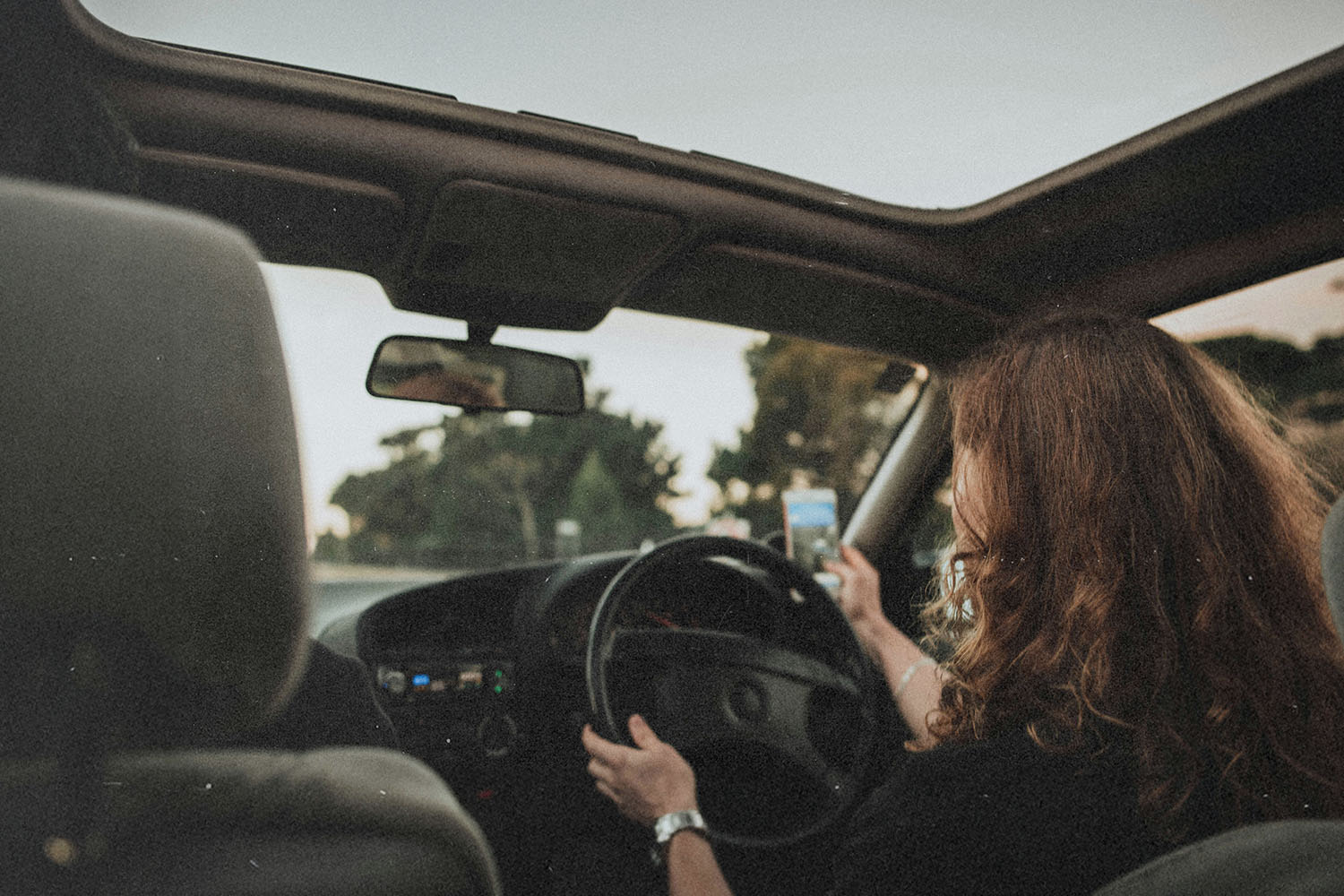 A person drives a car with the roof open, holding a phone in one hand while steering with the other