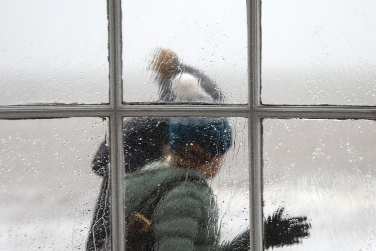Two people in winter clothing walk past a rain‑covered window, their figures softened by the water droplets on the glass