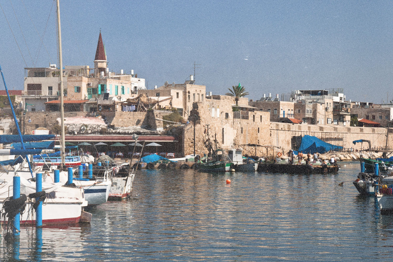 Boats and small fishing vessels float in a harbor beside stone buildings under a bright, clear sky