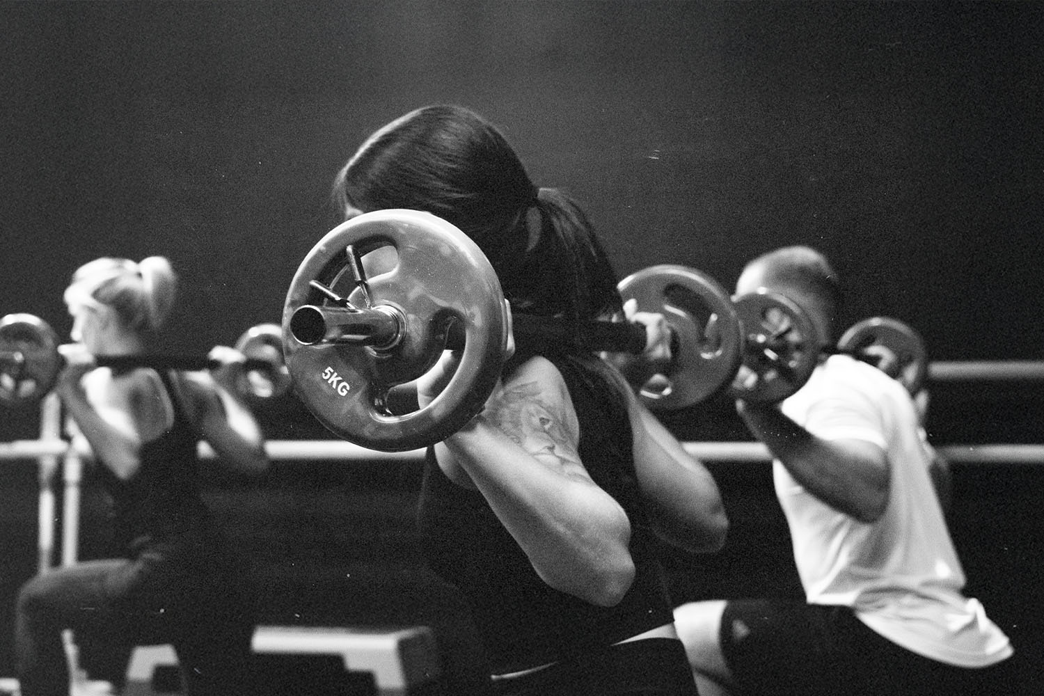 A group fitness class performs barbell squats, with participants lifting weighted bars across their shoulders