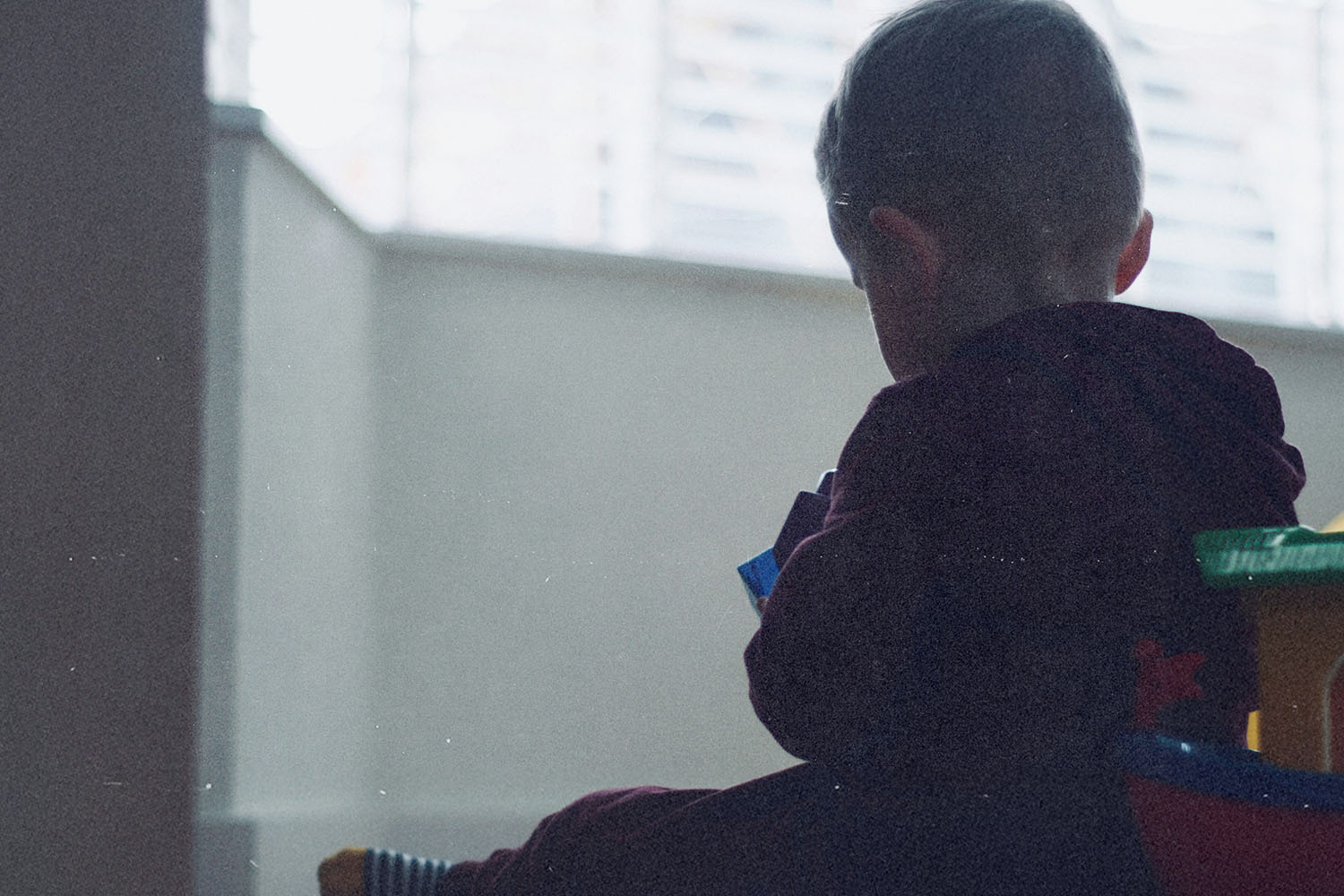 A young child sits indoors near a window, holding a toy while soft daylight filters through blinds behind them