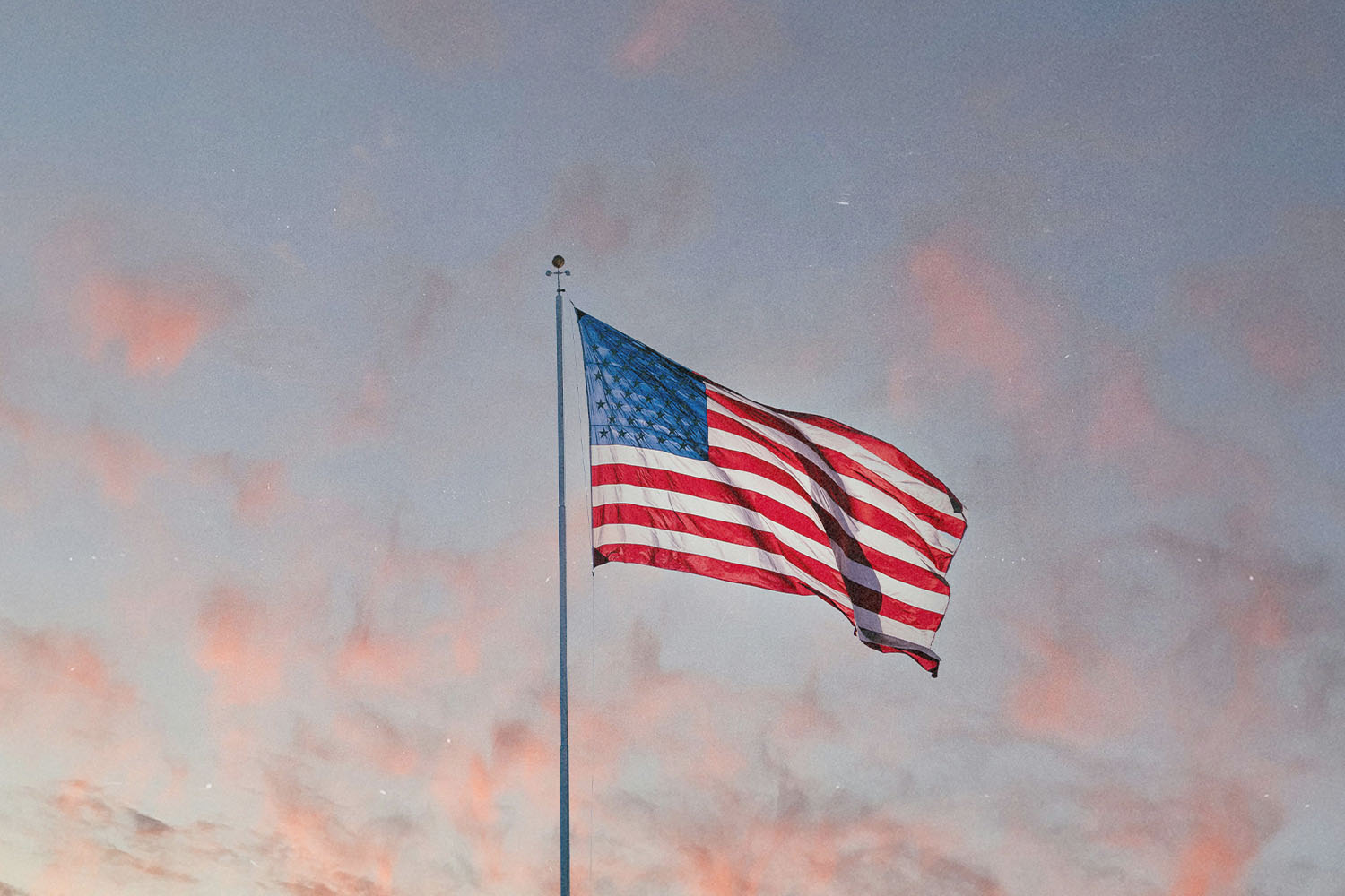 An American flag waves on a tall pole against a pastel evening sky with soft pink clouds