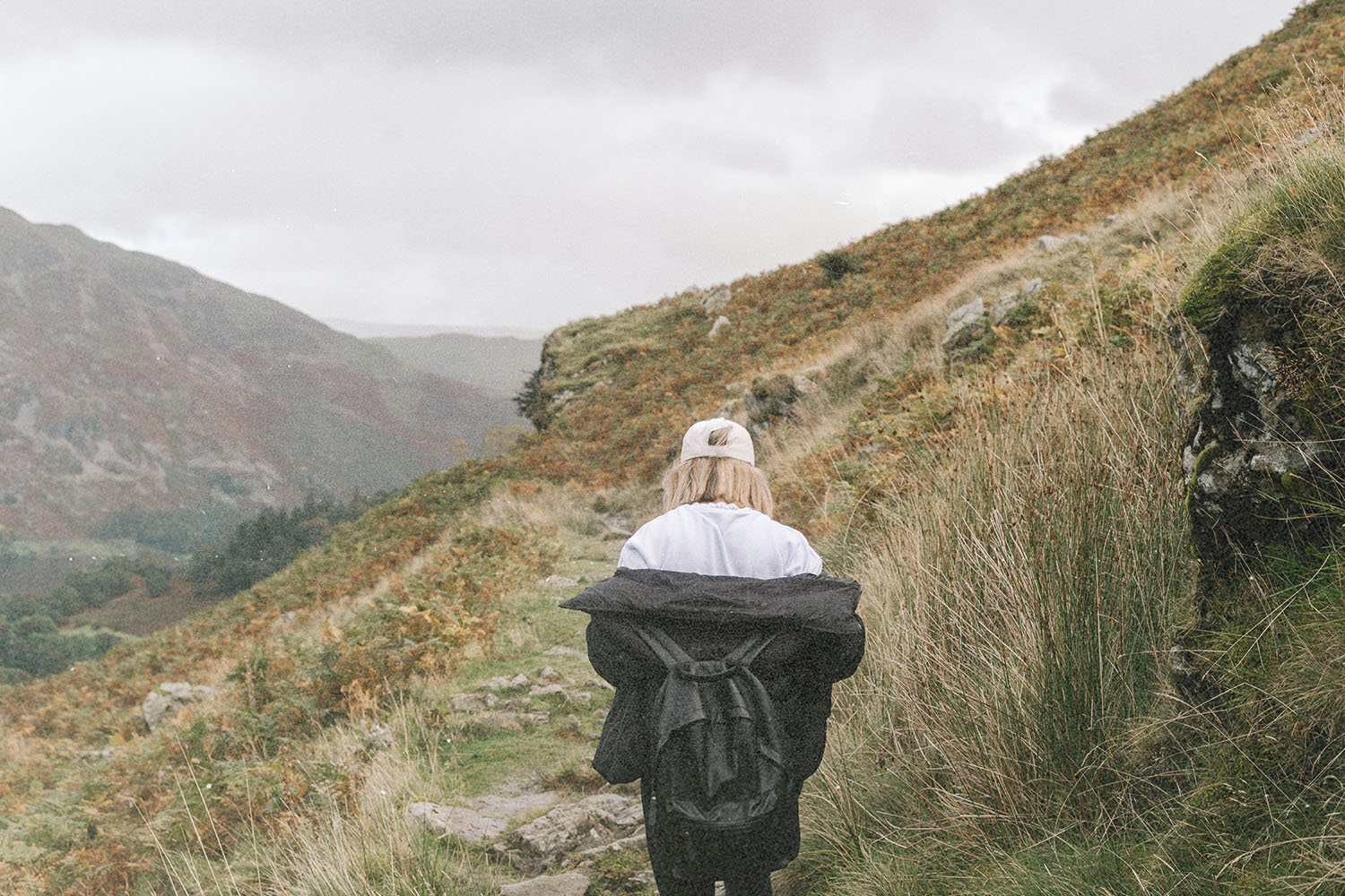 A person walks alone along a narrow mountain trail, wearing a light jacket and backpack as misty hills rise on both sides