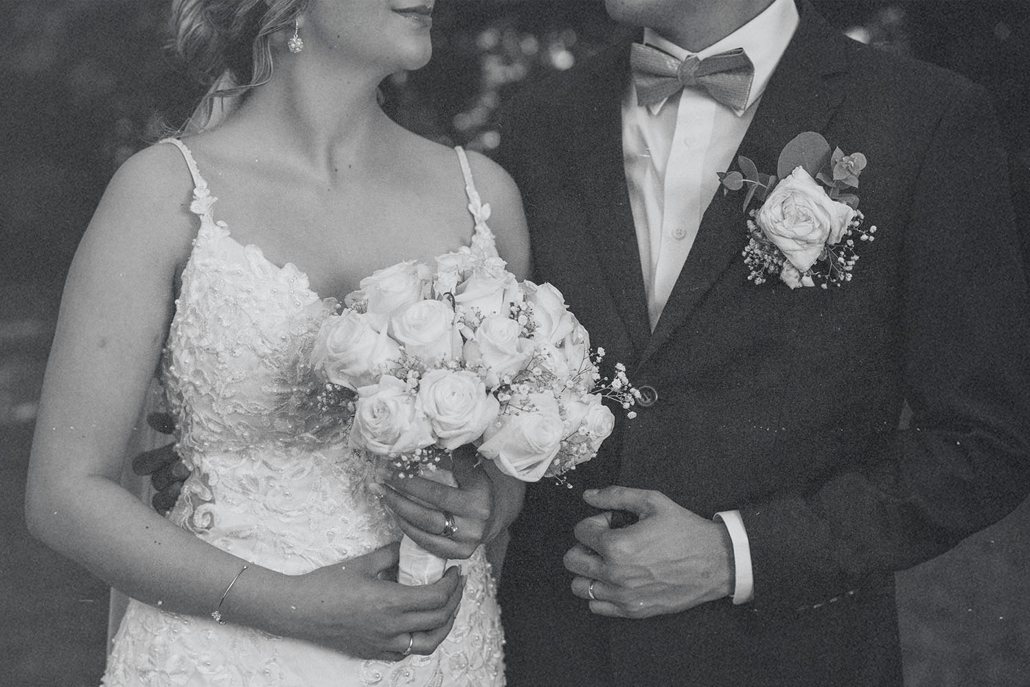 A bride and groom stand close together, the bride holding a bouquet of roses while the groom adjusts his suit jacket