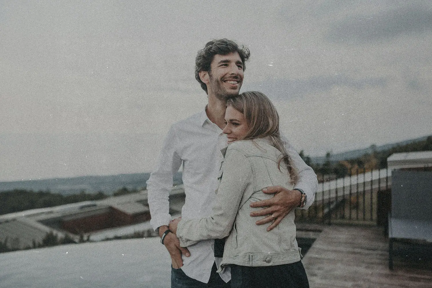 Two people stand together outdoors on a rooftop or terrace, with one person’s arm wrapped around the other as they look toward the landscape