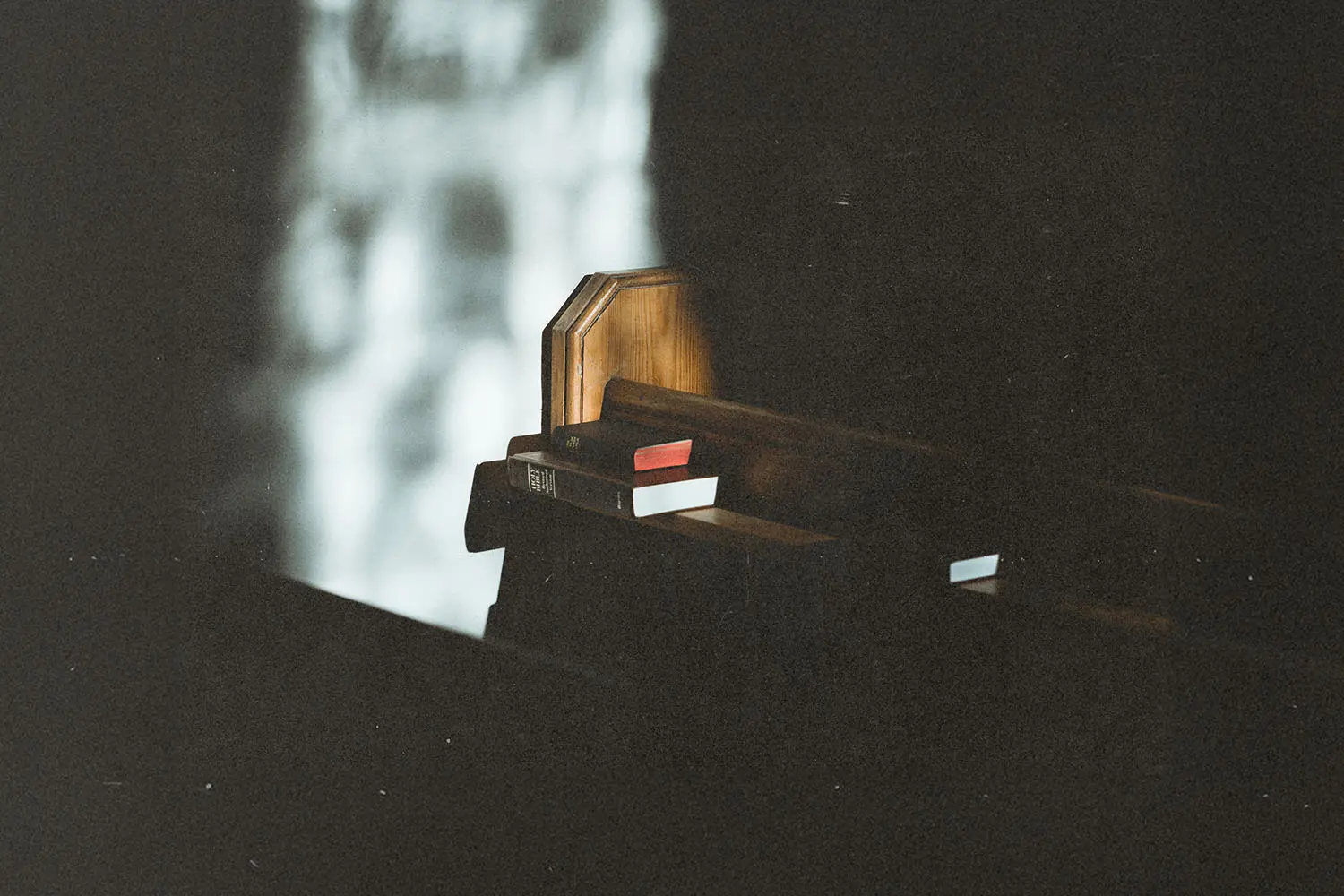 A wooden church pew is lit by a sliver of light, with hymnals and a Bible tucked into the rack behind it