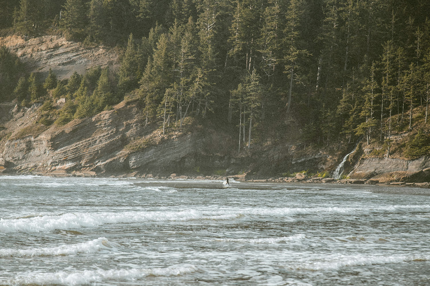 Gentle ocean waves roll toward a forested coastline with steep rocky cliffs and a small waterfall flowing into the sea
