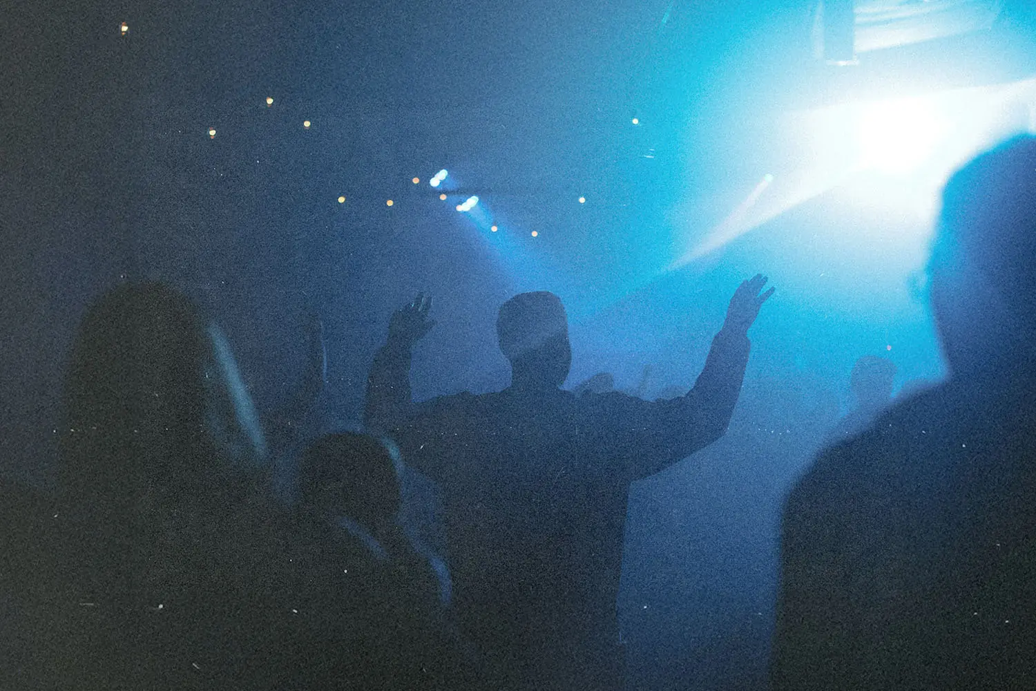 A silhouetted crowd stands in blue stage lighting, with one person raising both hands toward a bright spotlight