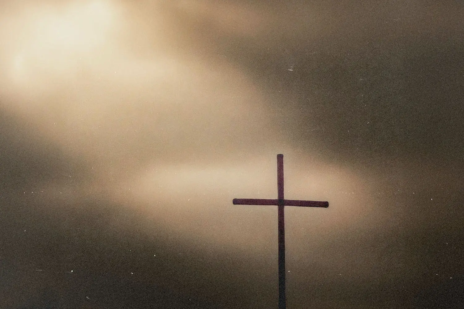A simple wooden cross stands silhouetted against a dramatic sky with a beam of light breaking through the clouds