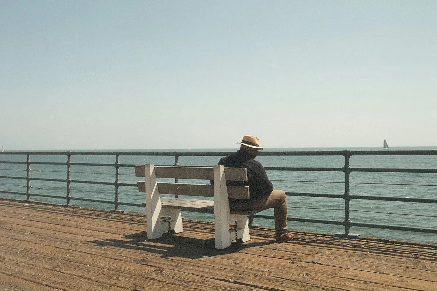A person wearing a hat sits alone on a wooden pier bench, looking out over pier railing at a calm ocean
