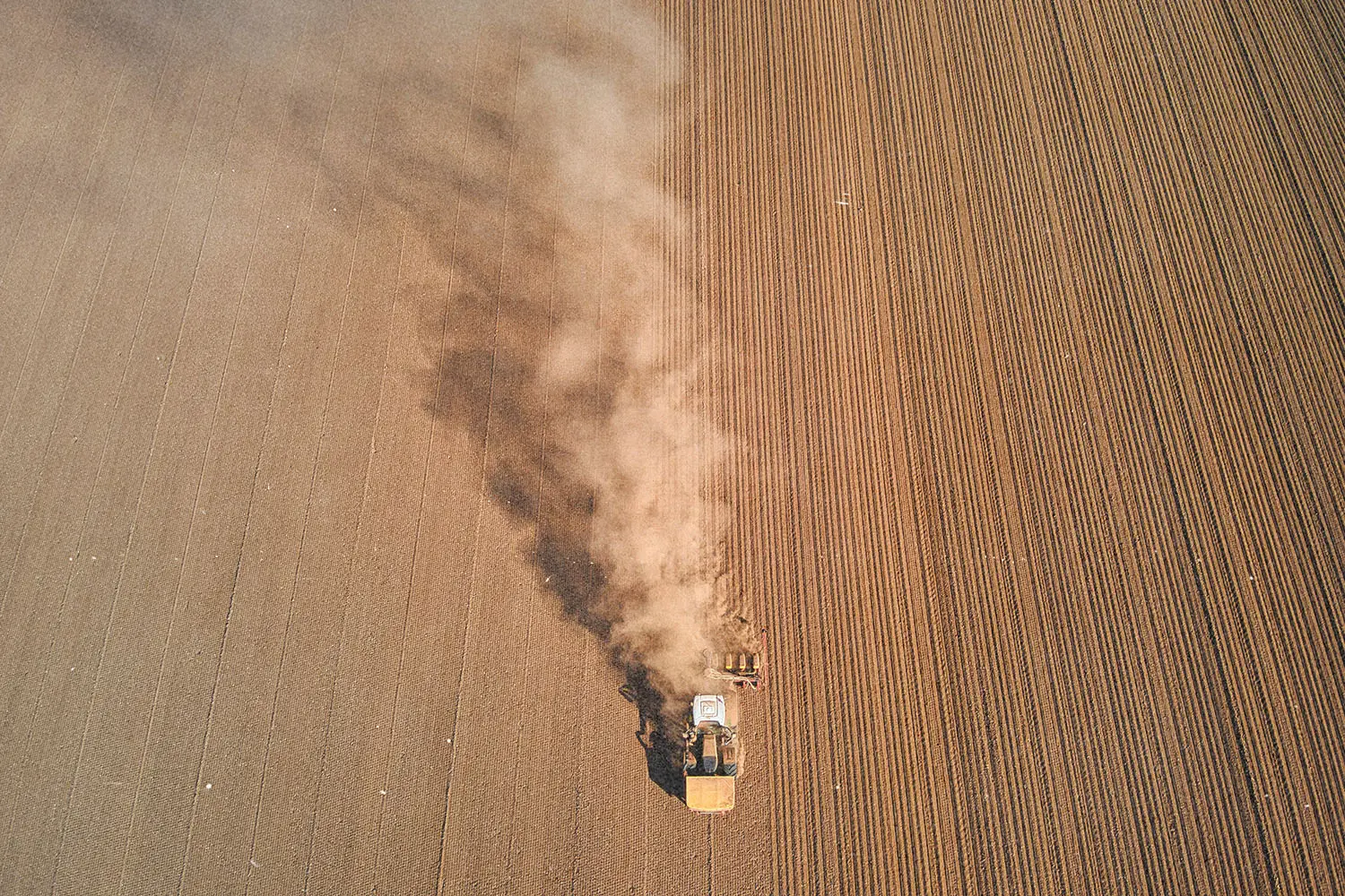 A tractor moves across a large plowed field, leaving a cloud of dust trailing behind it