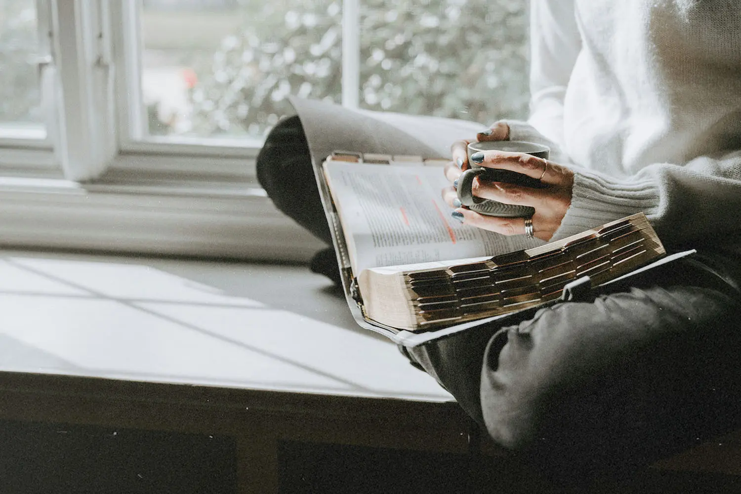 A person sits by a window holding a mug while reading an open Bible resting on their lap in soft natural light