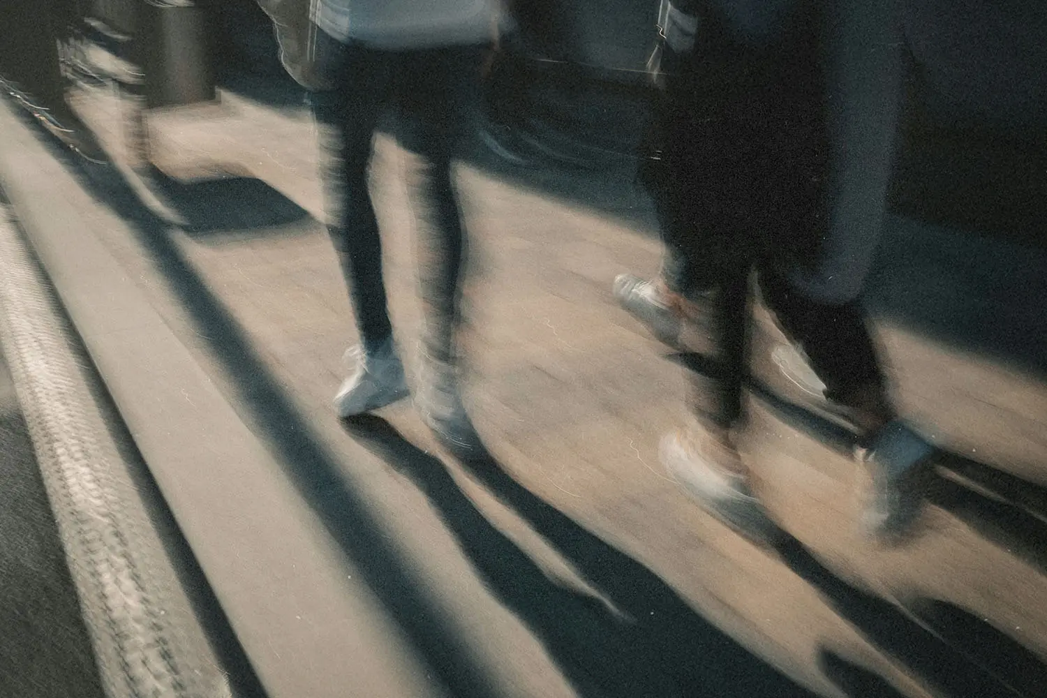 A motion‑blurred view of people walking along a sunlit sidewalk, their long shadows stretching across the ground