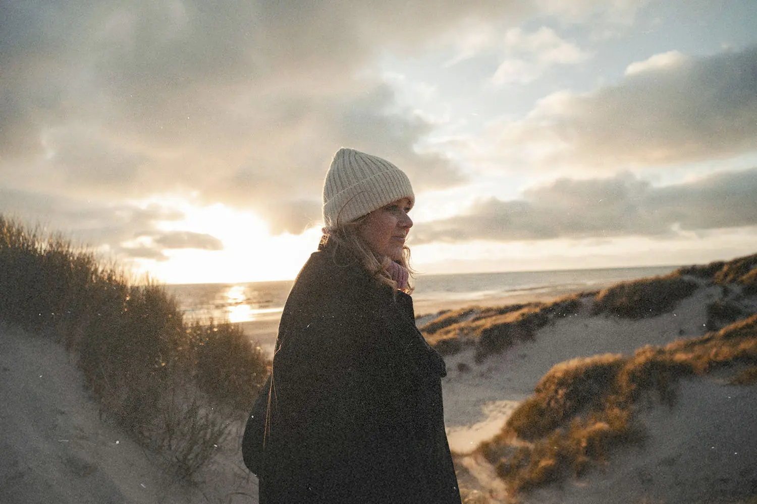 A woman wearing a knit beanie and coat walks along sandy dunes near the beach at sunset, with warm light shining through clouds over the ocean