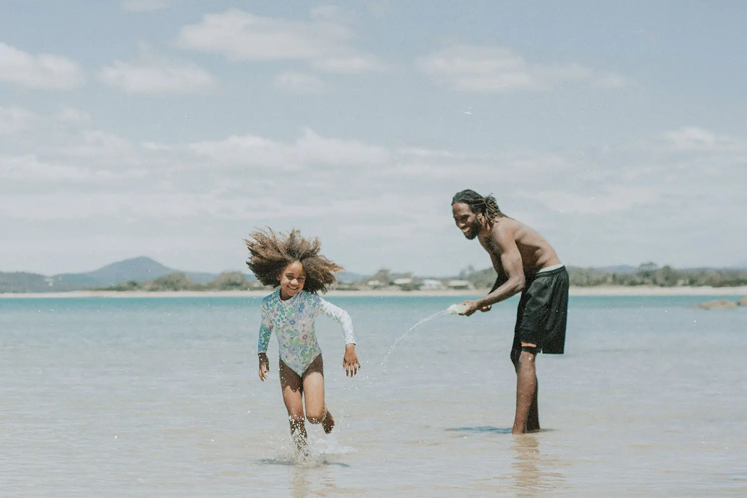 A child runs through shallow water at the beach as an adult playfully splashes water toward them under a bright sky