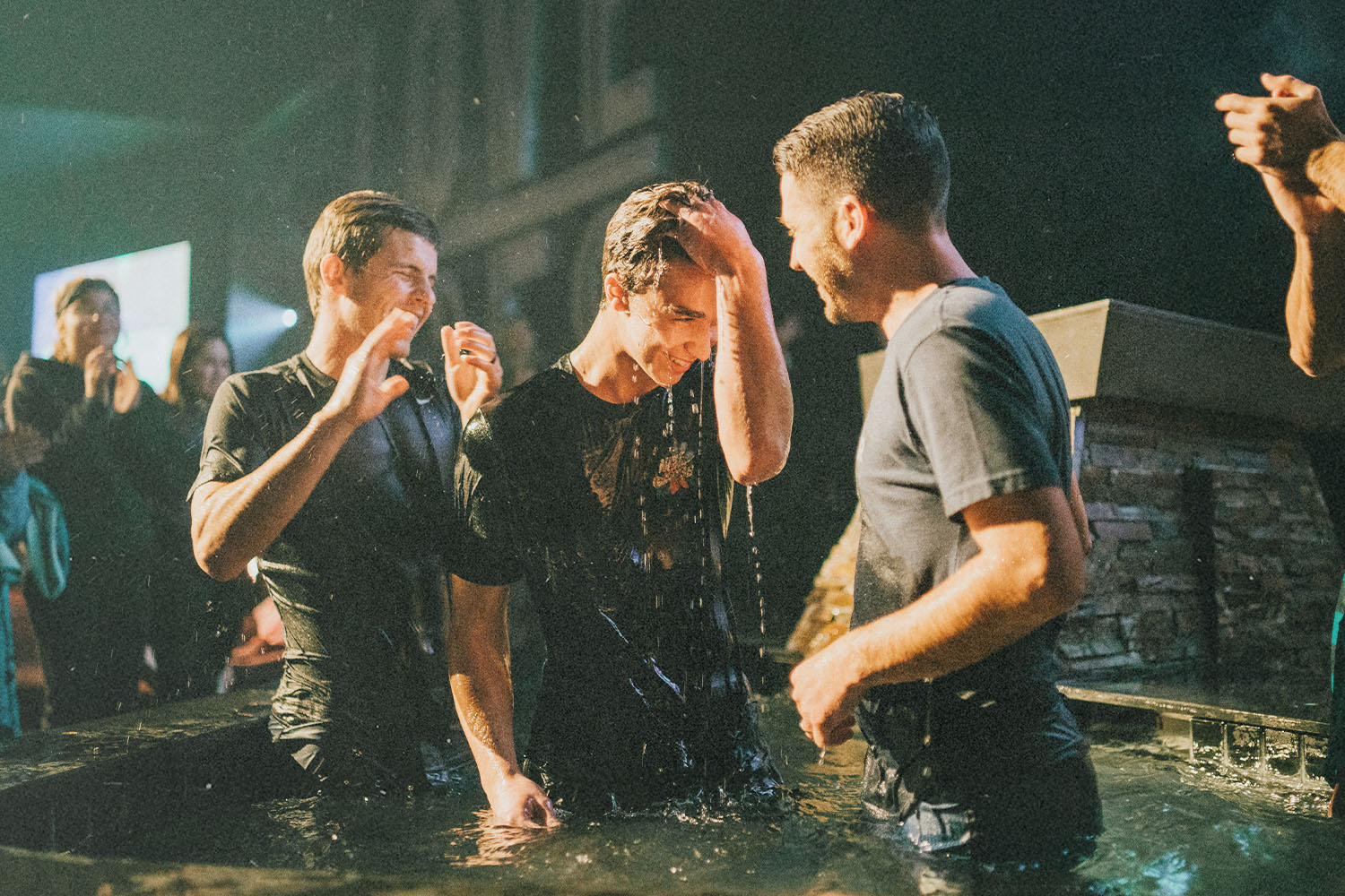 A group of people standing in a pool during a water baptism ceremony, with water splashing and onlookers nearby