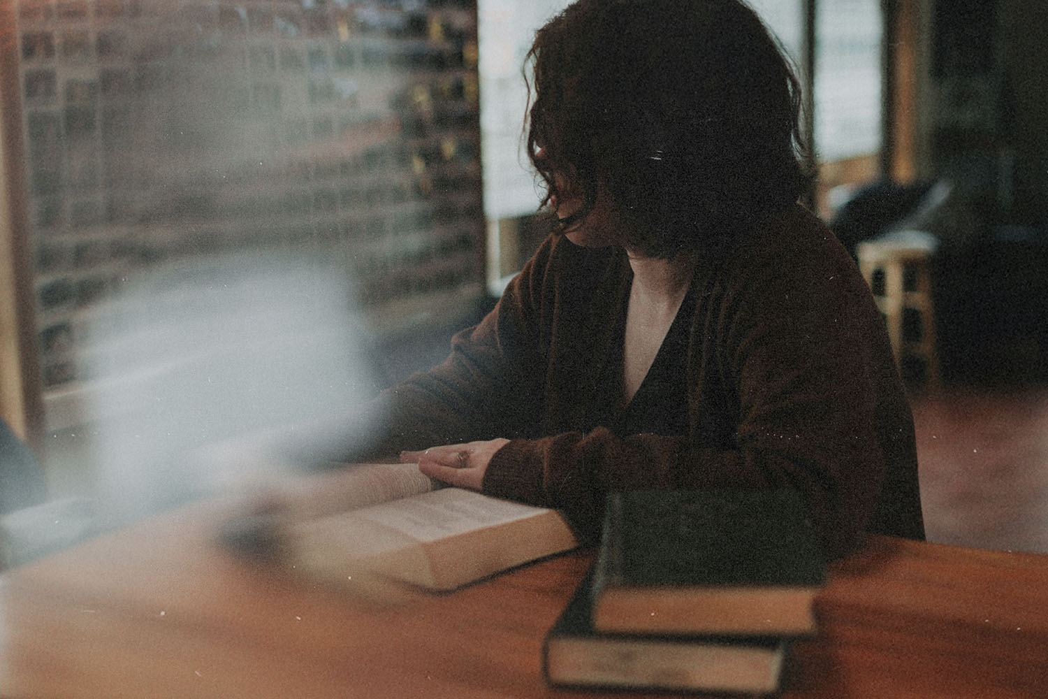 A person seated at a wooden table indoors with several books open and stacked nearby, in a softly lit room with brick walls