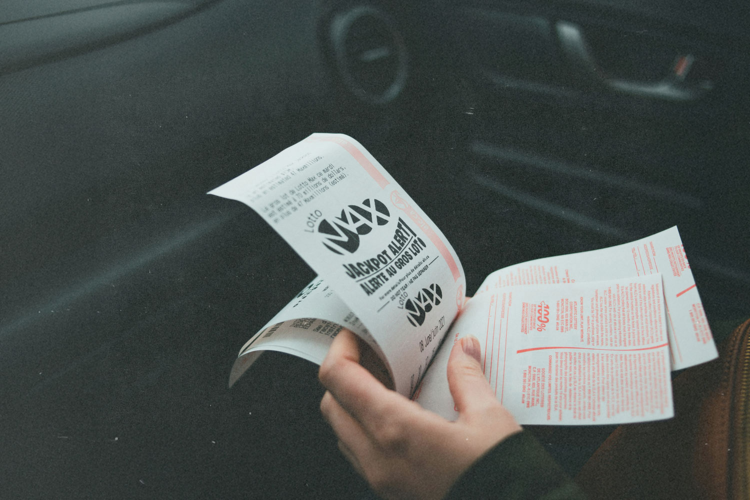 Hands hold printed lottery tickets inside a parked car, with the dashboard and interior visible in the background
