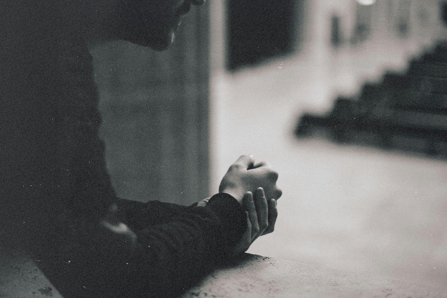 A person leans forward against a concrete ledge with hands clasped together, looking out over a quiet, empty walkway in a muted indoor setting