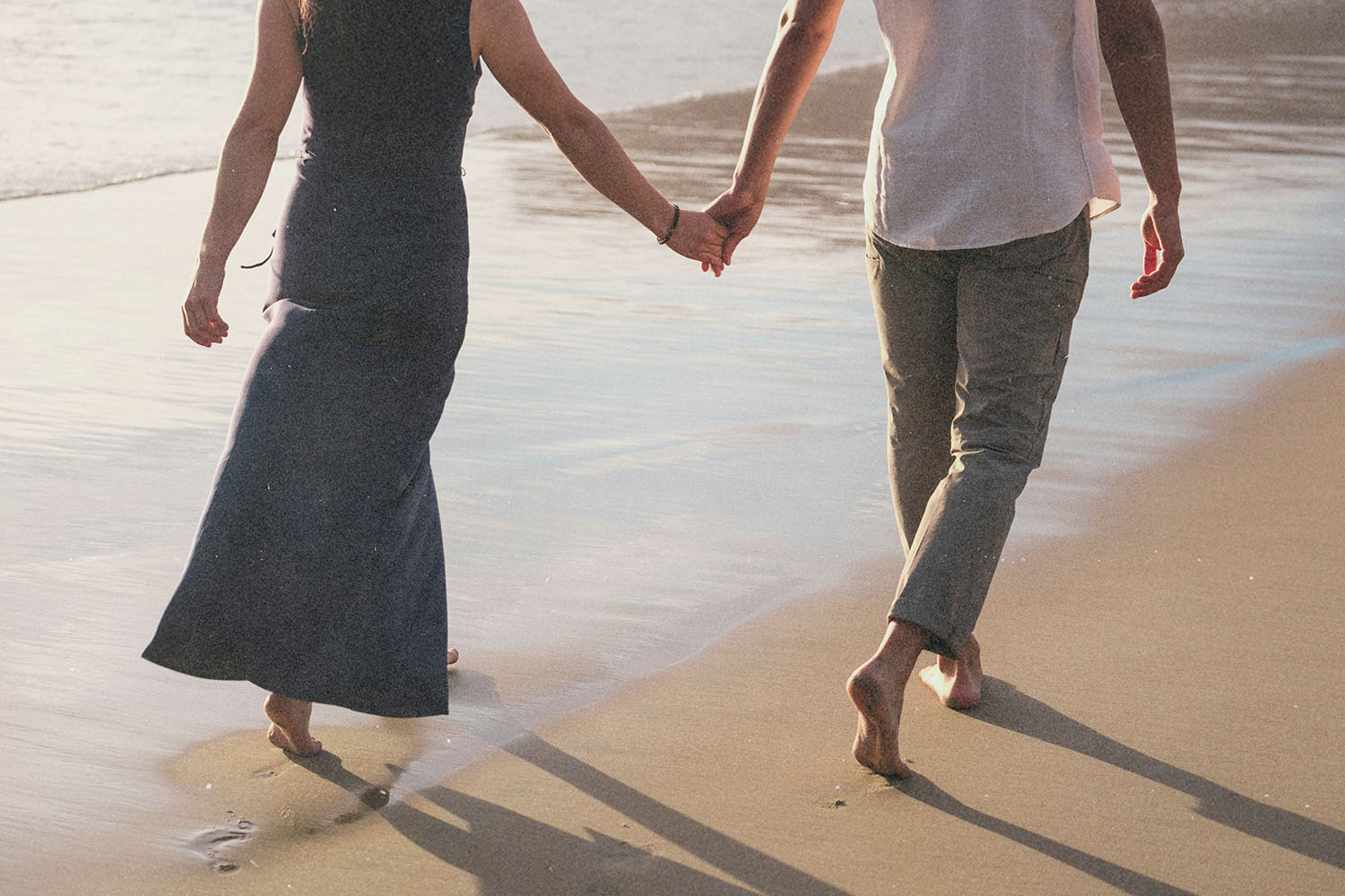 Two people walking hand in hand along a sandy shoreline where waves meet the beach