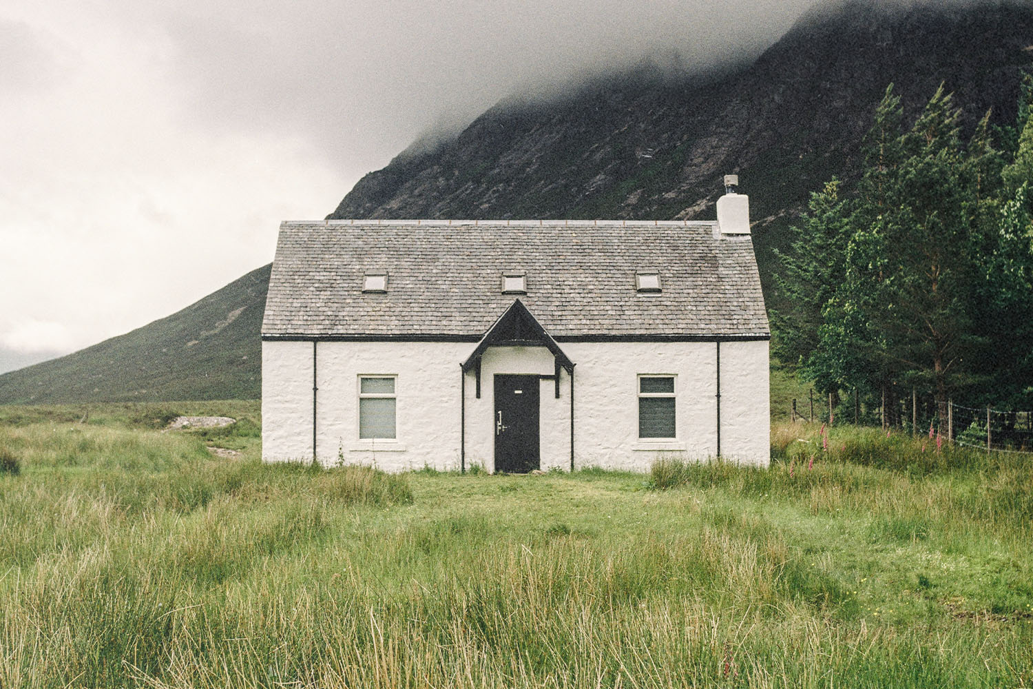 A small white house stands alone in a grassy field with misty mountains rising behind it