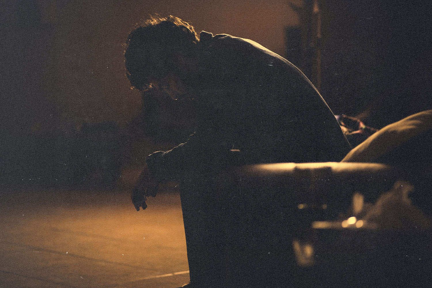 A person sits leaning forward on a couch in a dimly lit room, illuminated by warm, low light