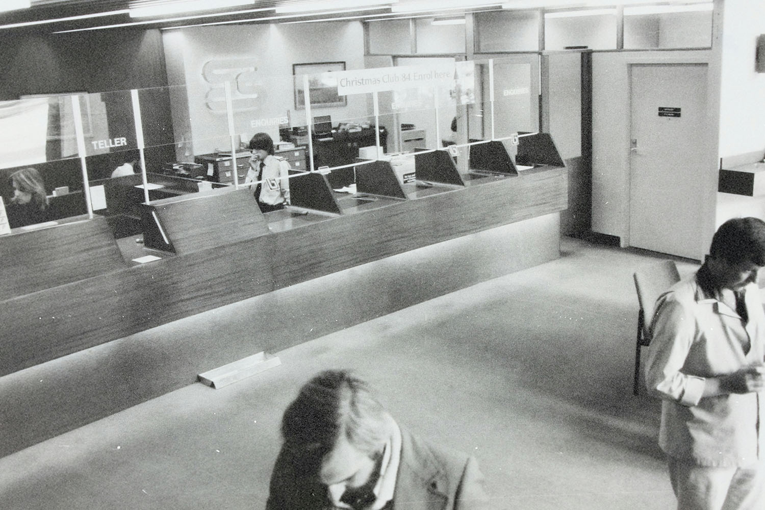 Interior of a bank lobby with teller counters, glass partitions, and several people moving through the space