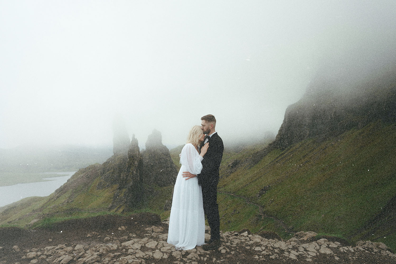 Newlywed couple stands close together on rocky ground, surrounded by misty green hills and dramatic cliffs