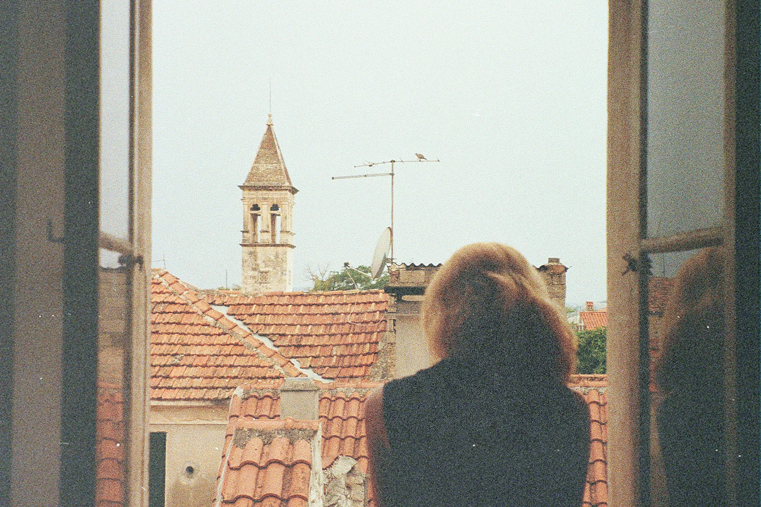 A person stands at an open window, looking out over red-tiled rooftops and a distant church tower