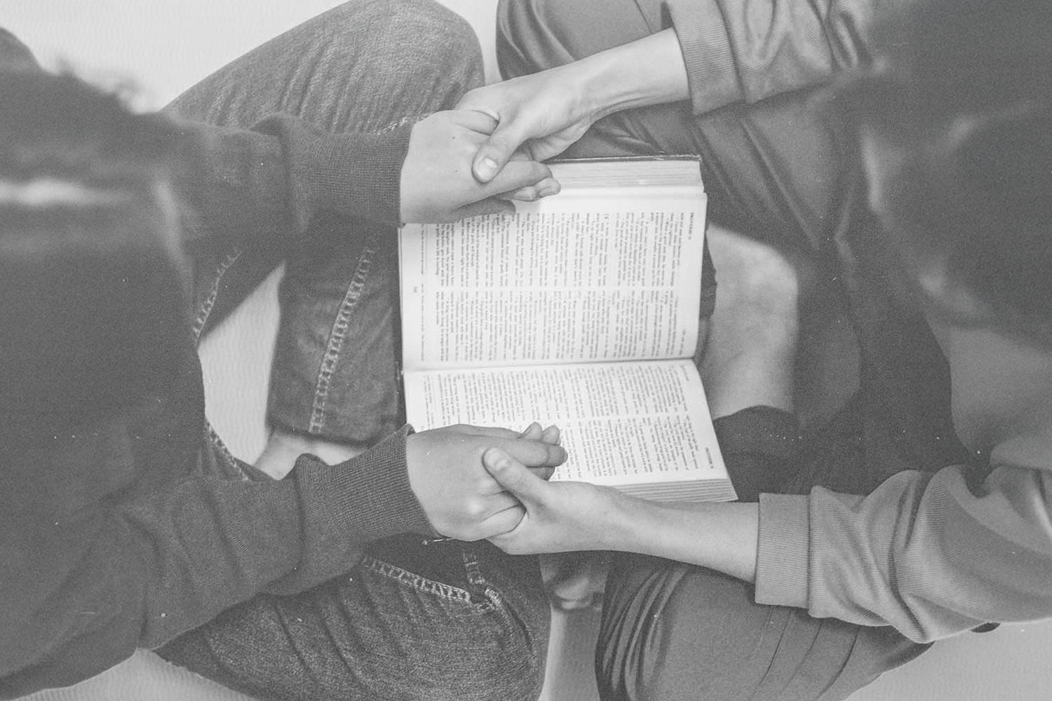 Two people sitting cross-legged, holding hands over an open book placed between them
