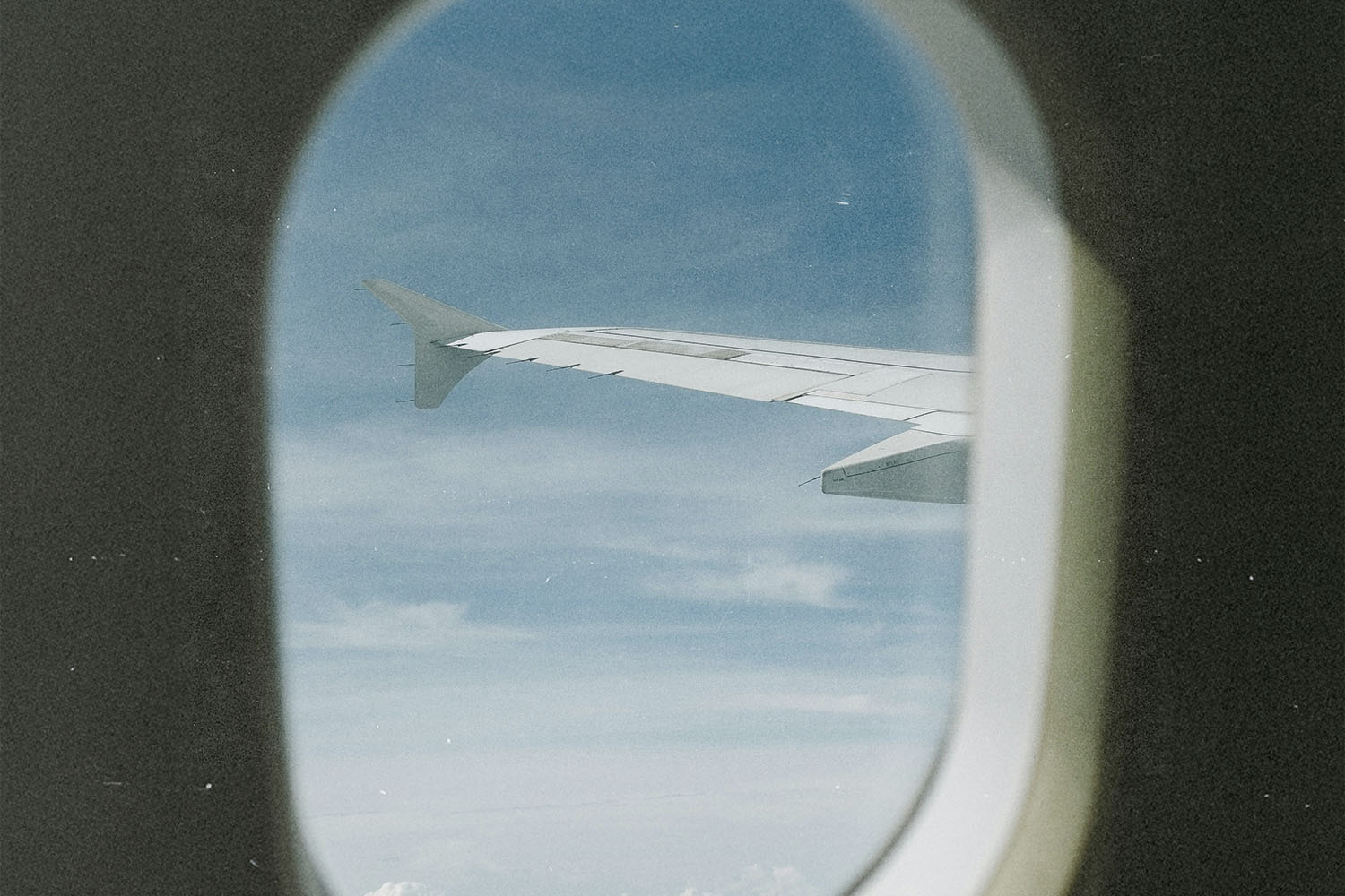 View from inside an airplane looking out a window at a wing against a blue sky with light clouds