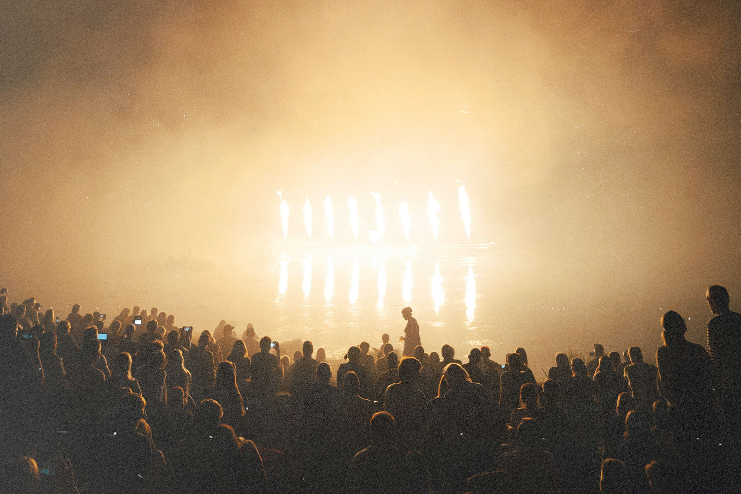 A large crowd gathered indoors, silhouetted against intense golden stage lights shining through haze