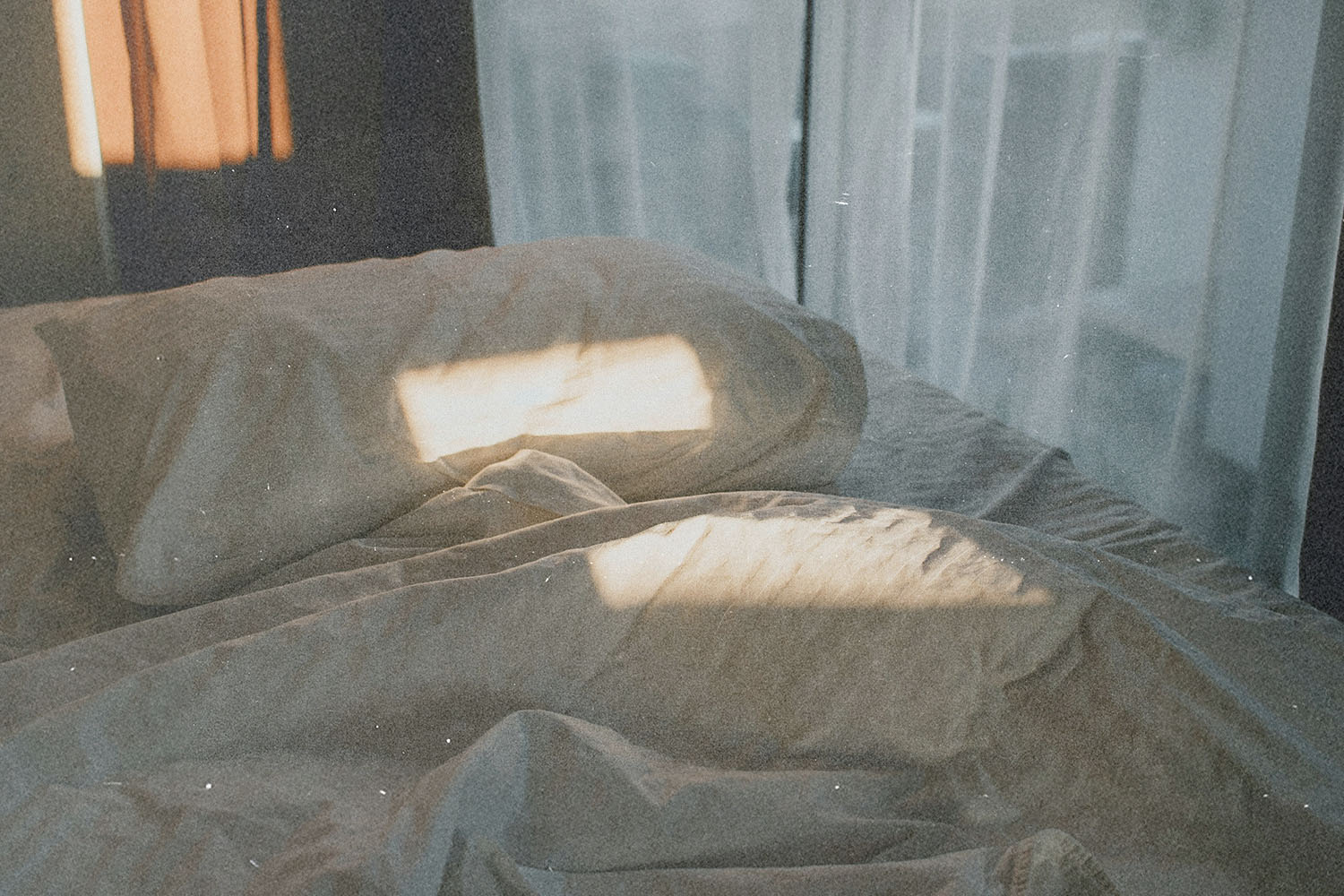 Soft morning light shining across rumpled bedding and pillows near a window with sheer curtains