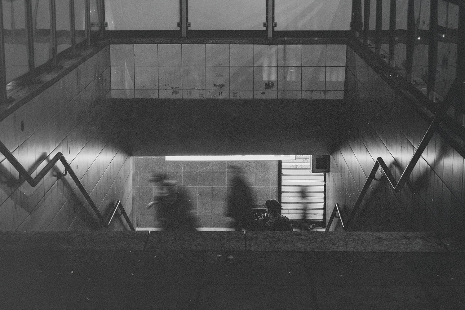 Dimly lit subway entrance with blurred figures descending toward the platform and tiled walls along the stairs