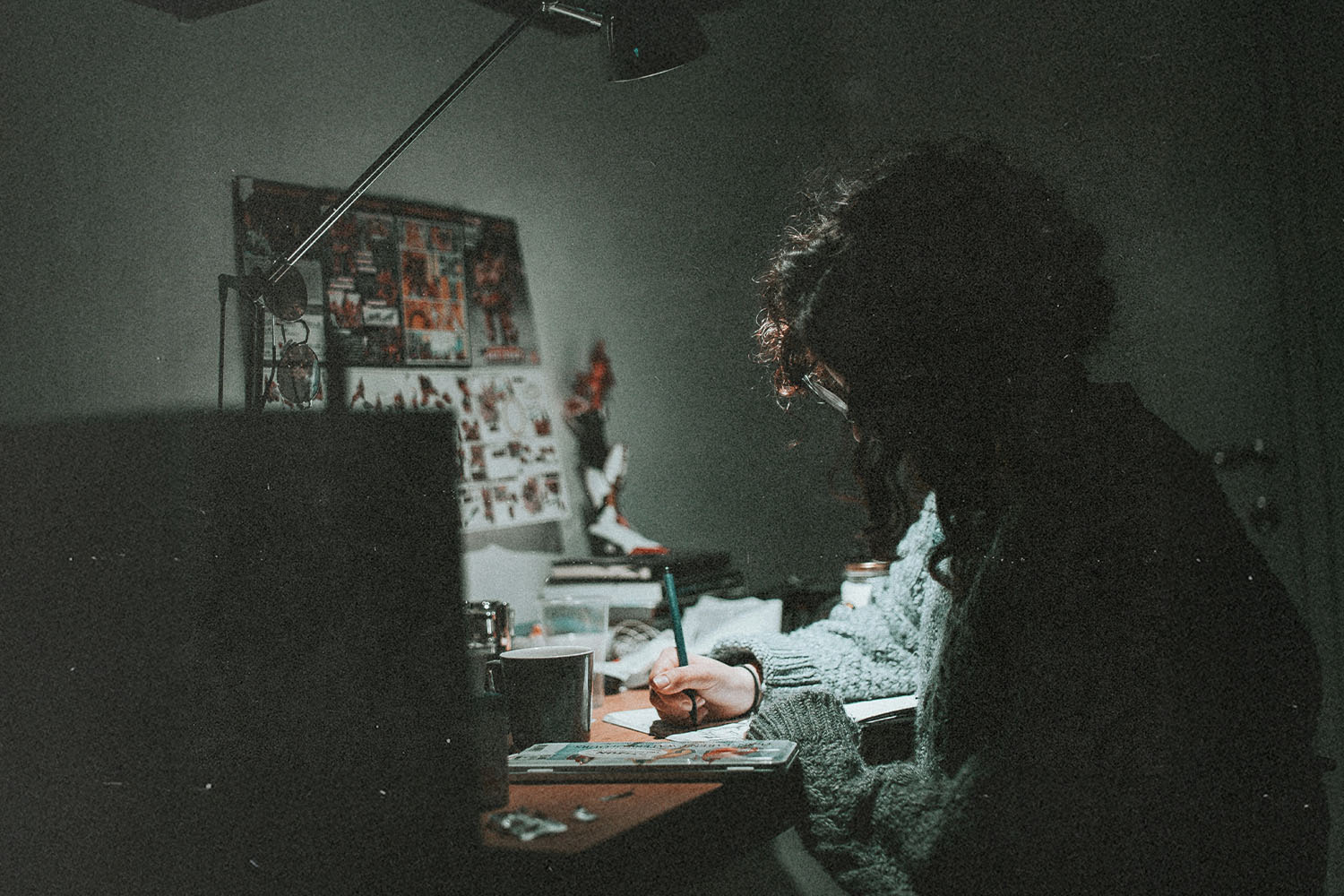 Person seated at a cluttered desk writing under a focused desk lamp in a dimly lit room