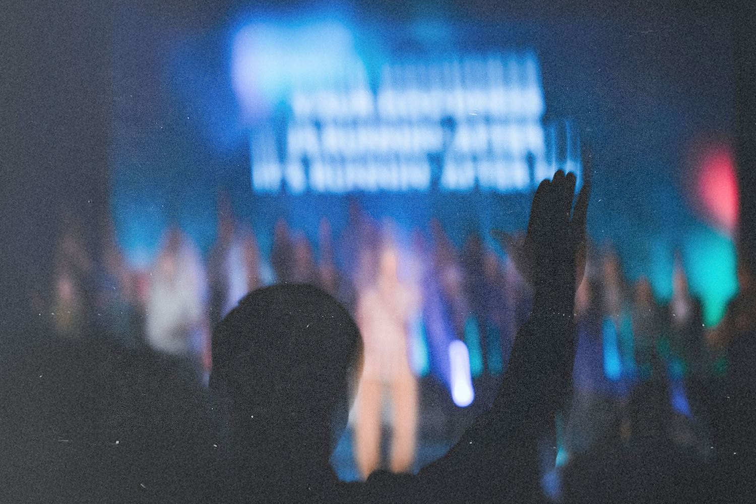 Silhouetted figure raising a hand in a crowded worship gathering with colorful stage lights in the background
