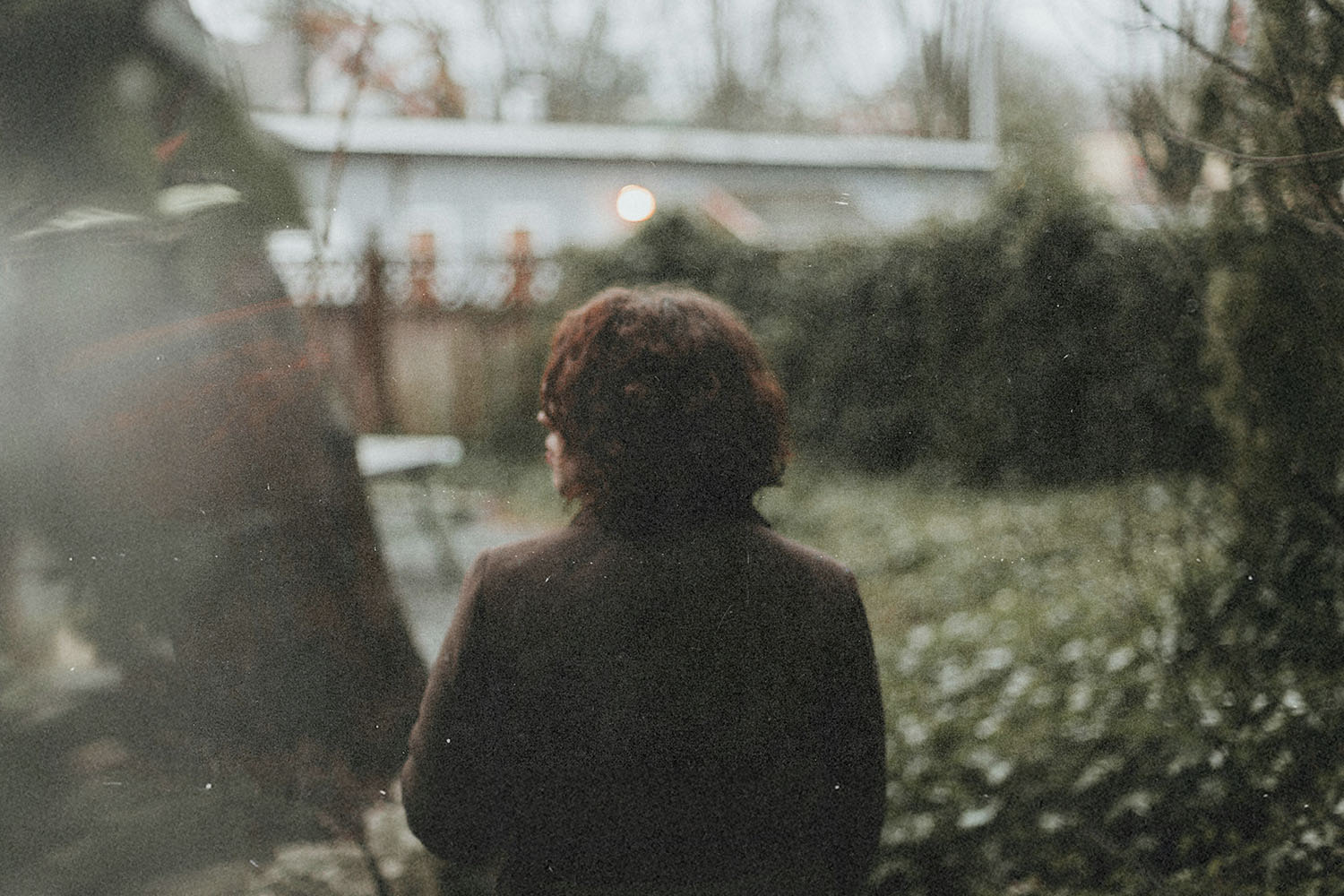 Person standing in a garden facing away from the camera, surrounded by trees and shrubs