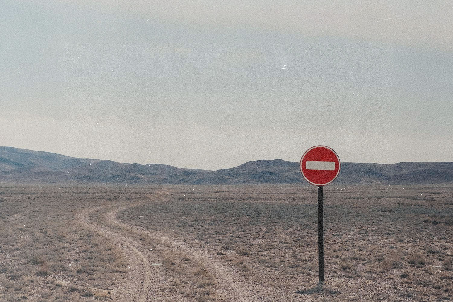 Red “do not enter” road sign standing beside a winding dirt track in an empty desert landscape