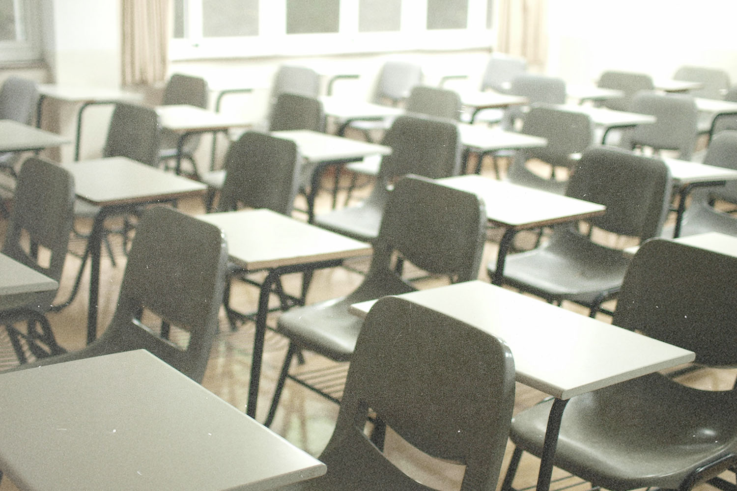 Rows of empty gray student desks in a brightly lit classroom with large windows