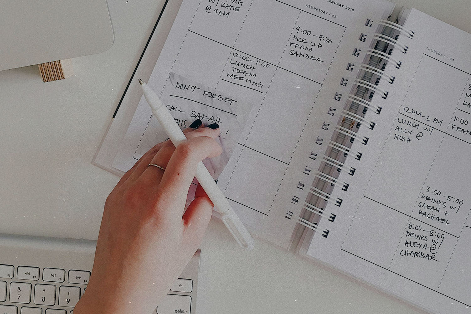 Hand holding a pen above an open planner next to a keyboard on a white desk