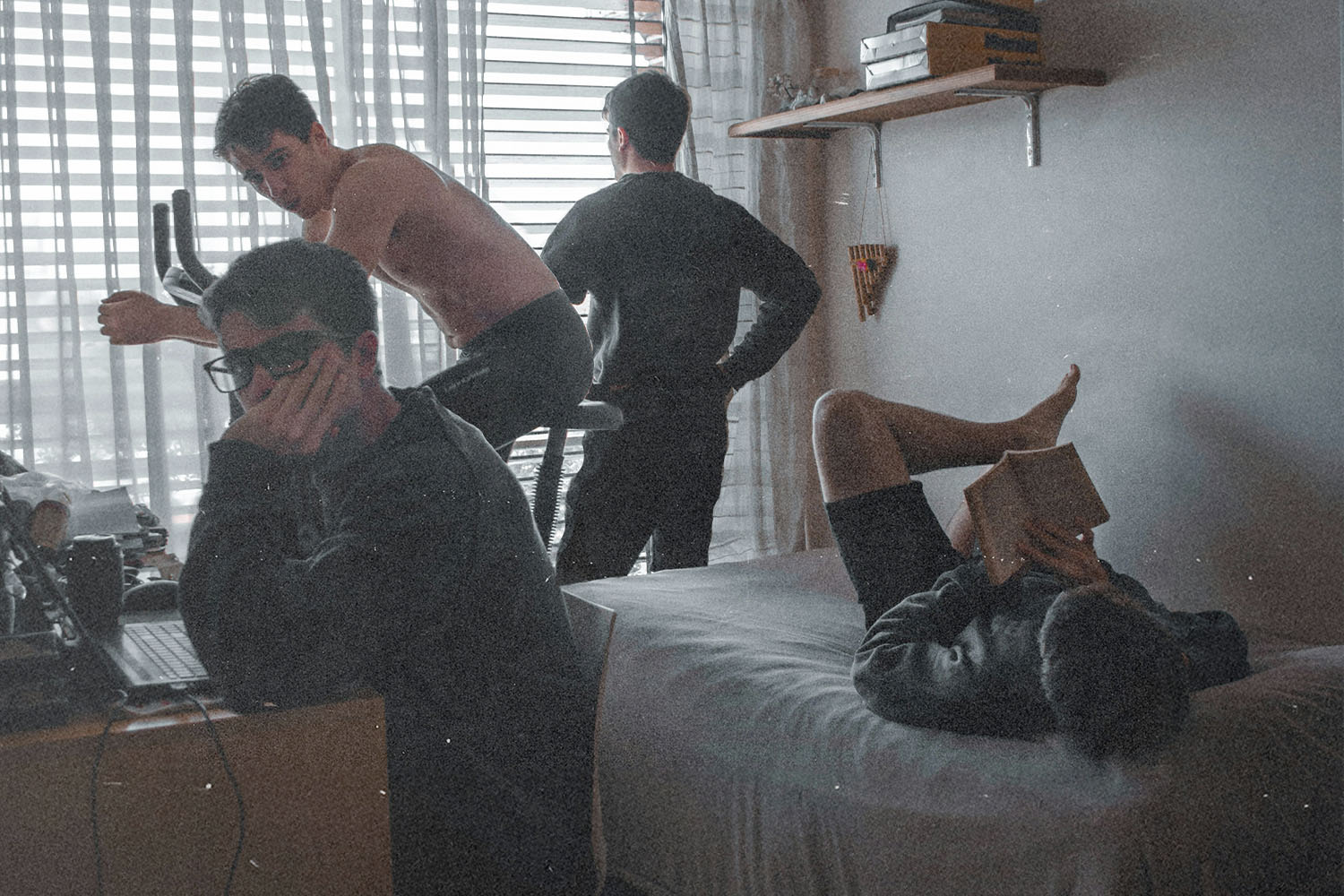 Four people in a small bedroom, one using a laptop, one on an exercise bike, one standing by the window, and one lying on a bed reading a book